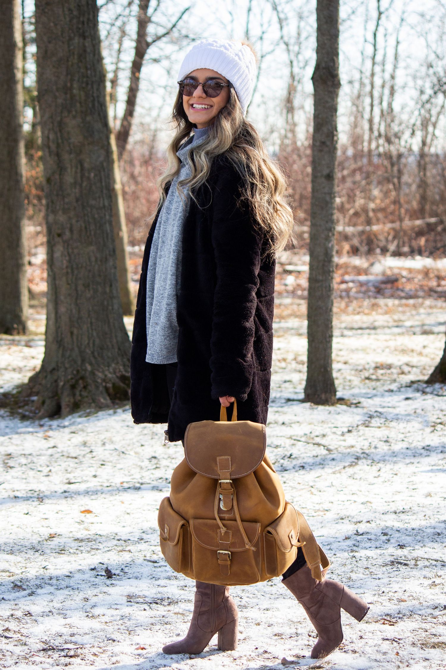 Toronto Portrait Photographer - Winter fashion portrait of person in black coat and beanie with brown leather backpack in snowy woods.