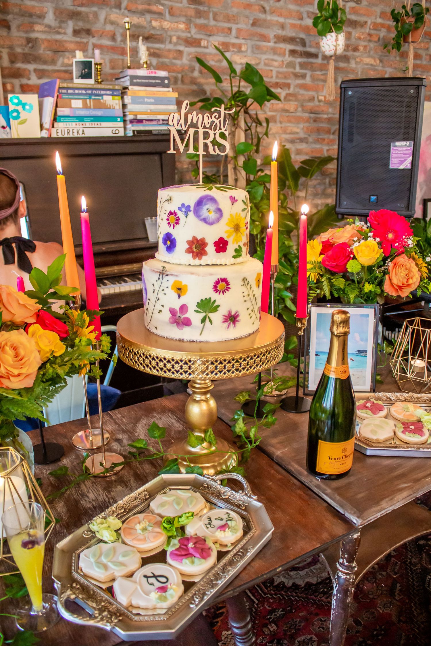 Toronto Event - Elegant wedding dessert display with tiered white cake, roses, and candles on rustic wooden table with exposed brick wall.