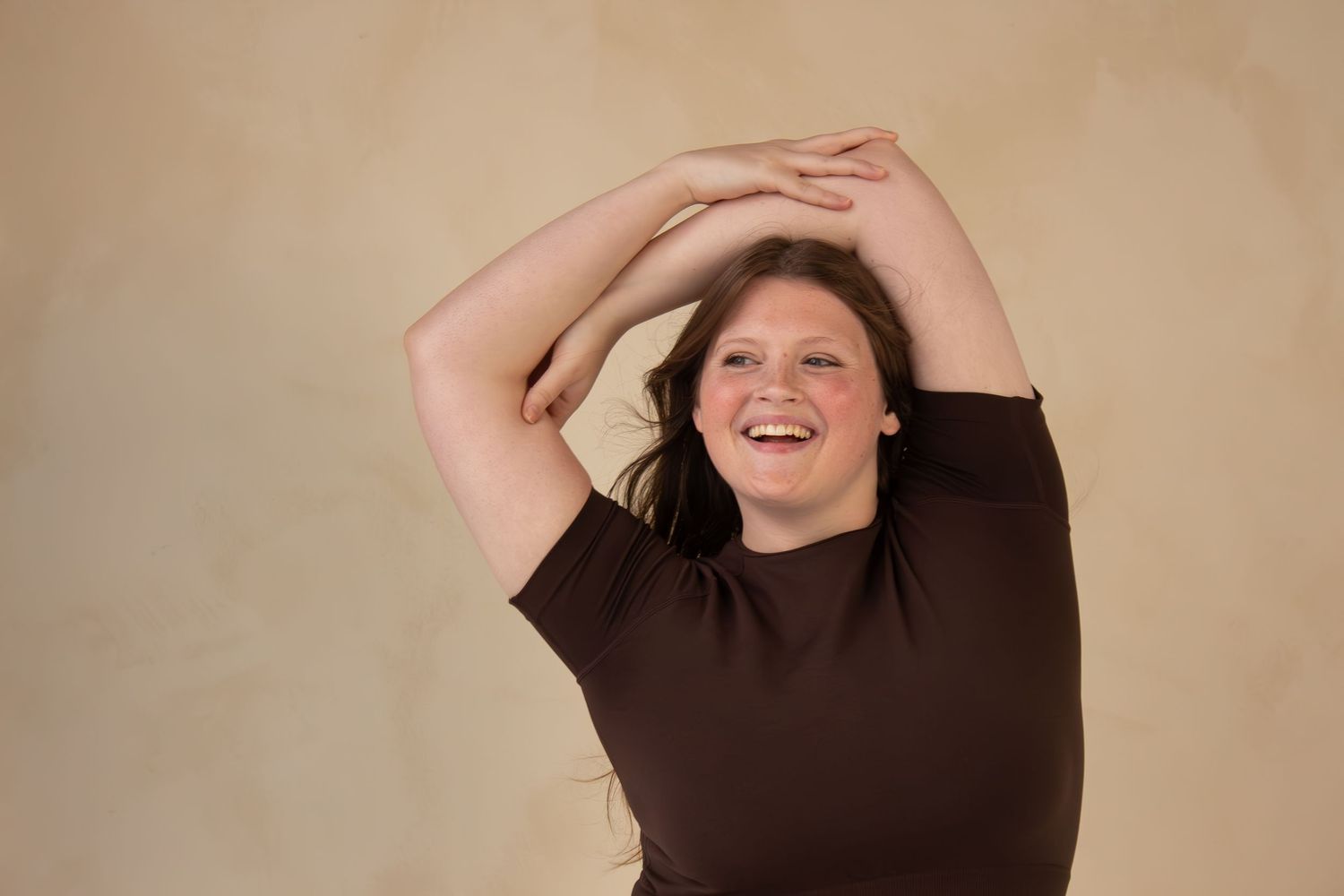 A person wearing a brown sweater poses playfully with their arms raised overhead against a beige background.
