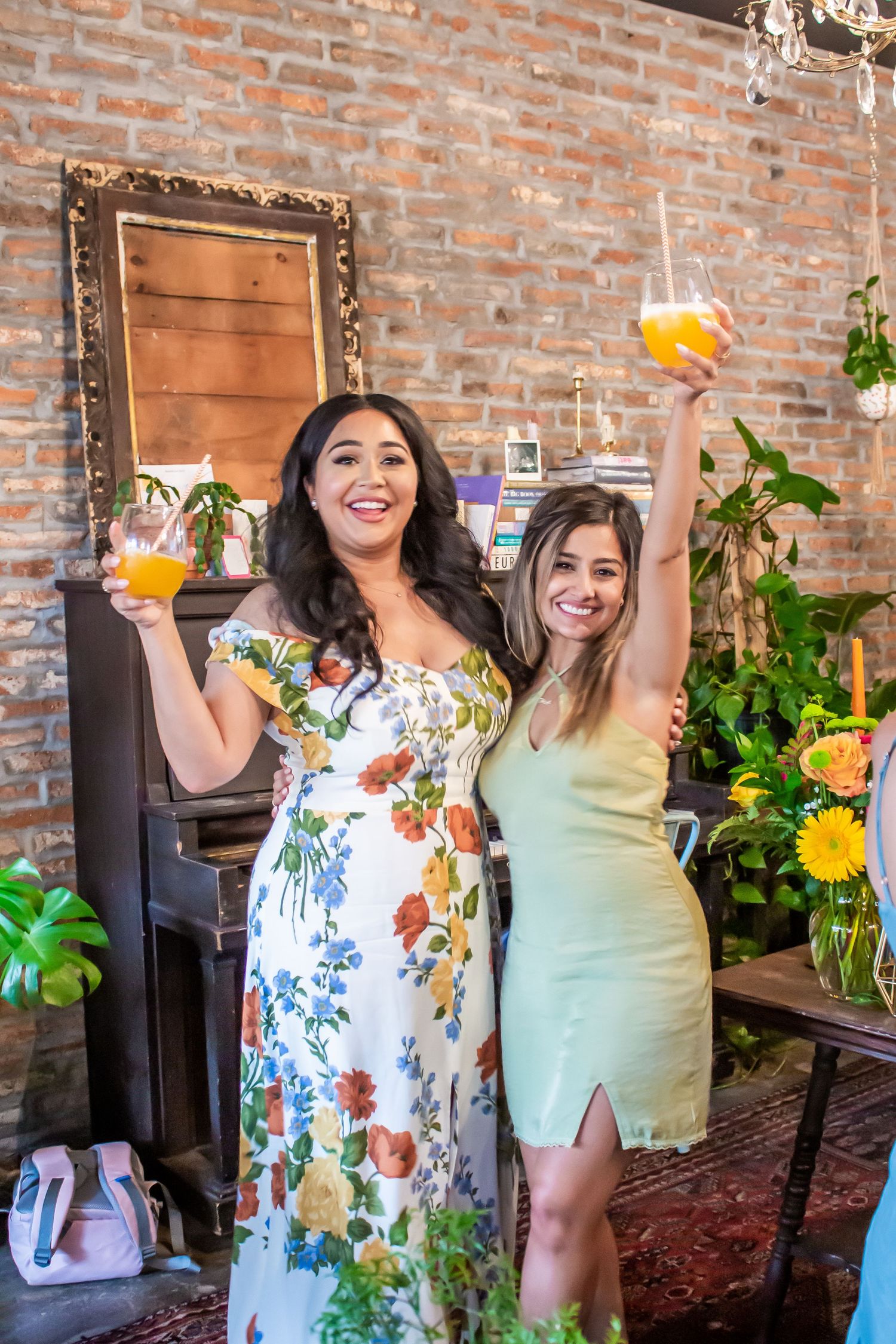 Two friends celebrate with colorful cocktails in front of an exposed brick wall with plants and decor.