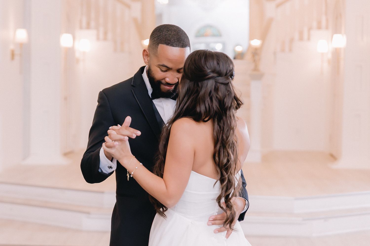 Couple dances intimately in an elegant white ballroom with soft lighting.