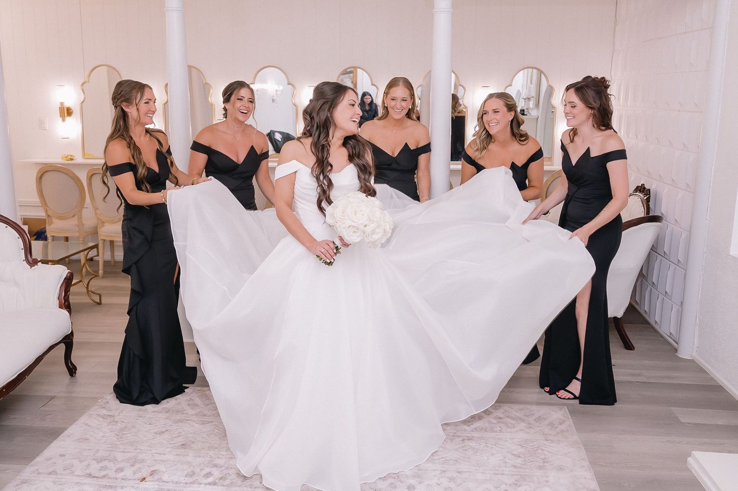 Bridesmaids in black dresses help arrange bride's white gown in bright hallway.