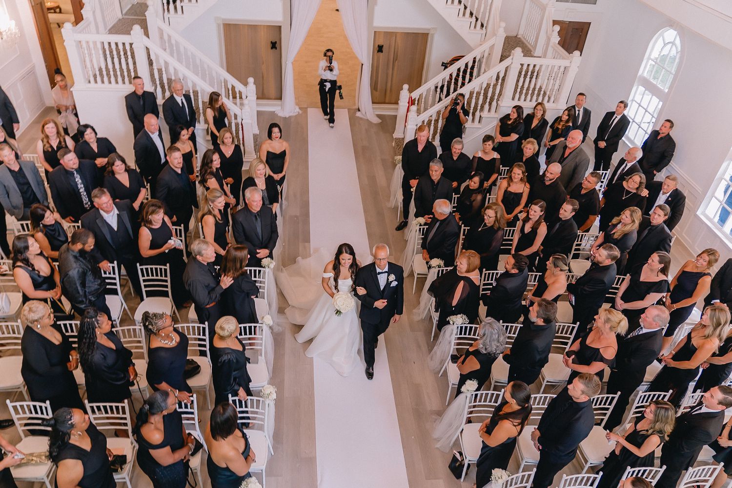 Large wedding party poses together in formal black attire inside bright white venue.