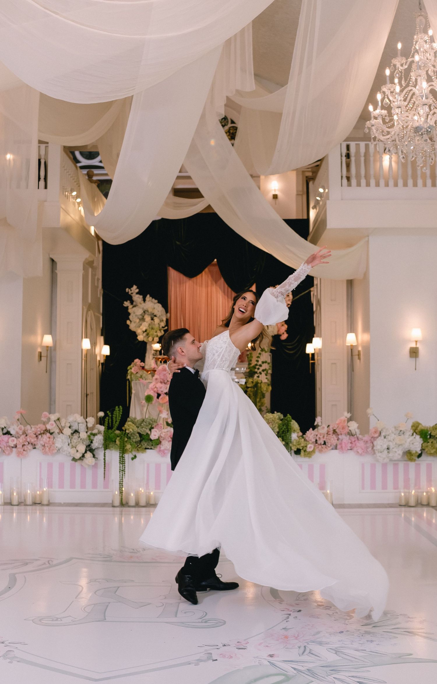 Bride in white gown lifted by groom on elegant dance floor with floral decor and draped ceiling at wedding reception.