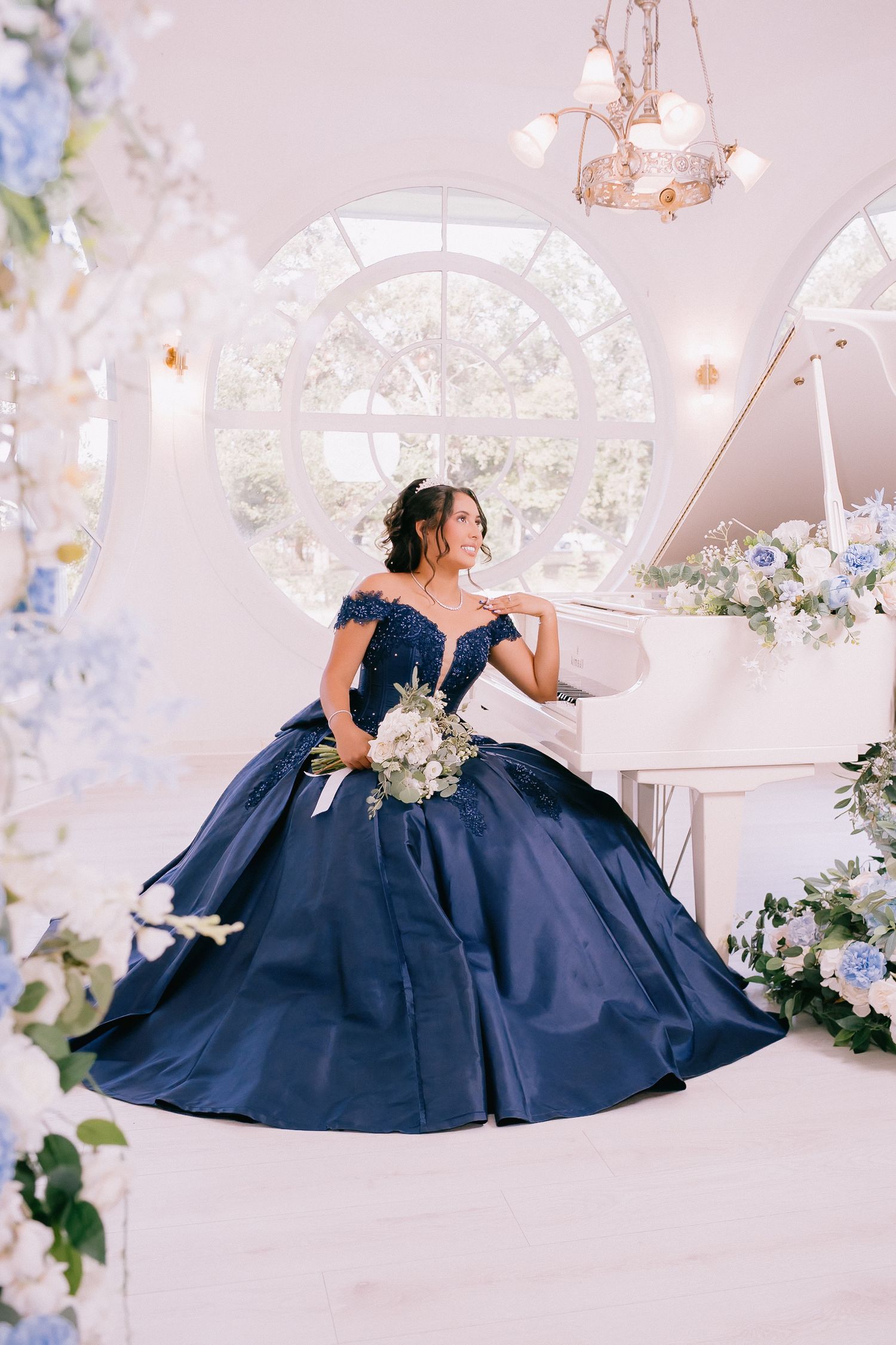 Dreamy portrait in navy ball gown beside white piano adorned with blue and white floral arrangements.