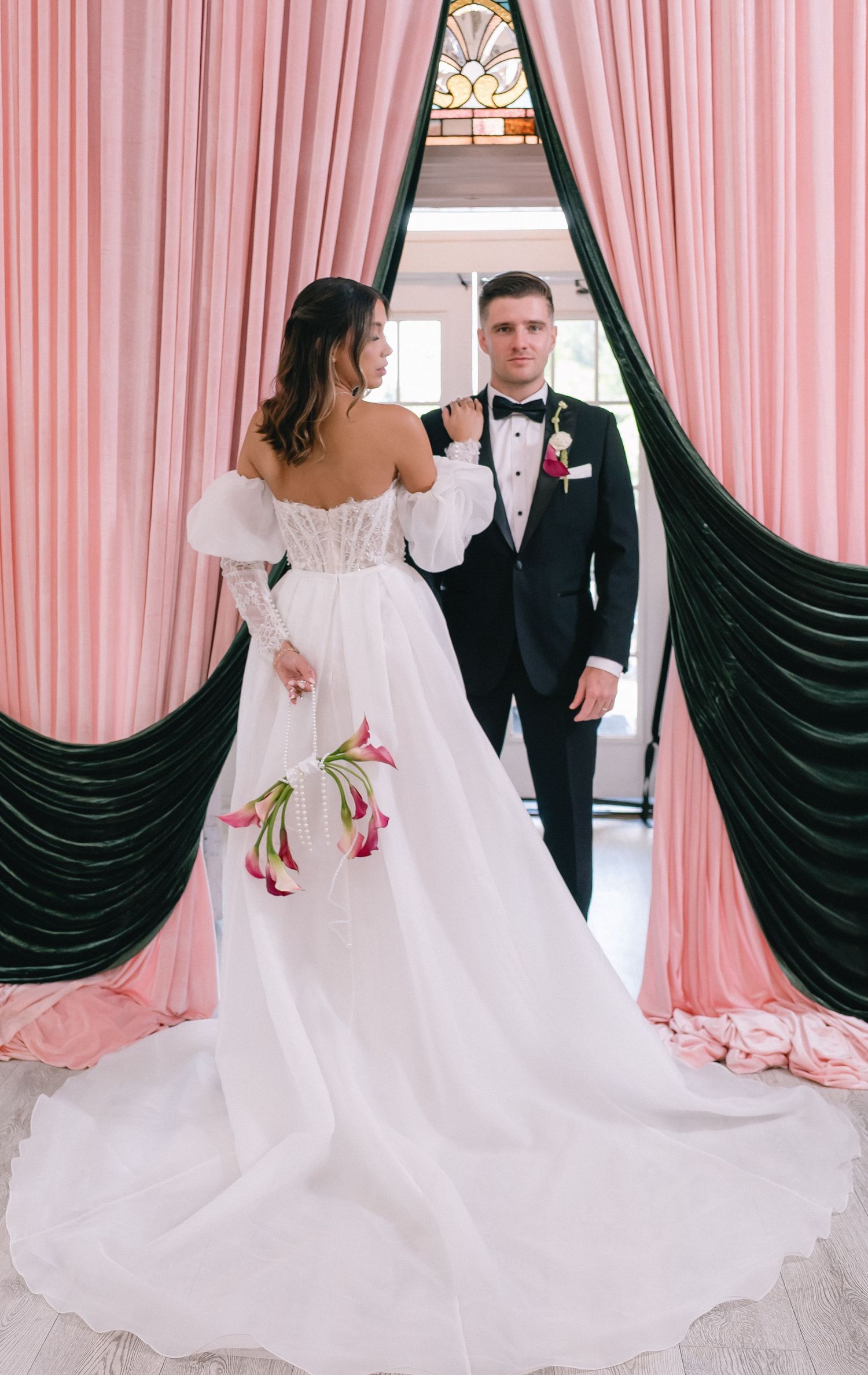 Bride in off-shoulder white gown and groom in black tuxedo pose between pink and green draping curtains.