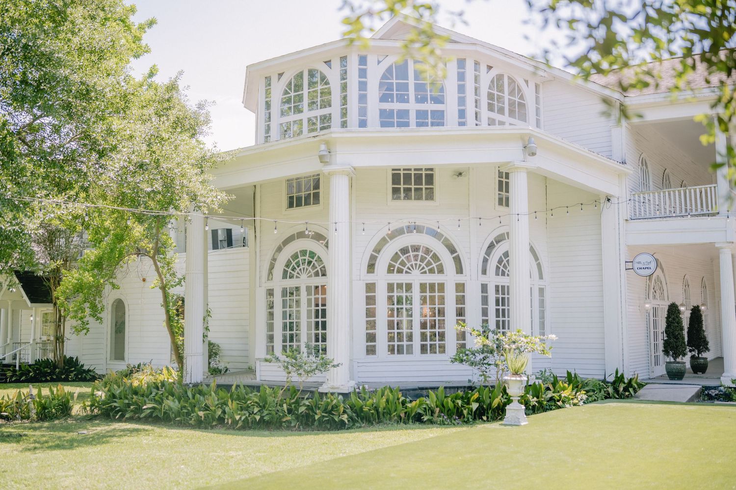 Elegant white mansion exterior with arched windows and lush landscaping on a sunny day.