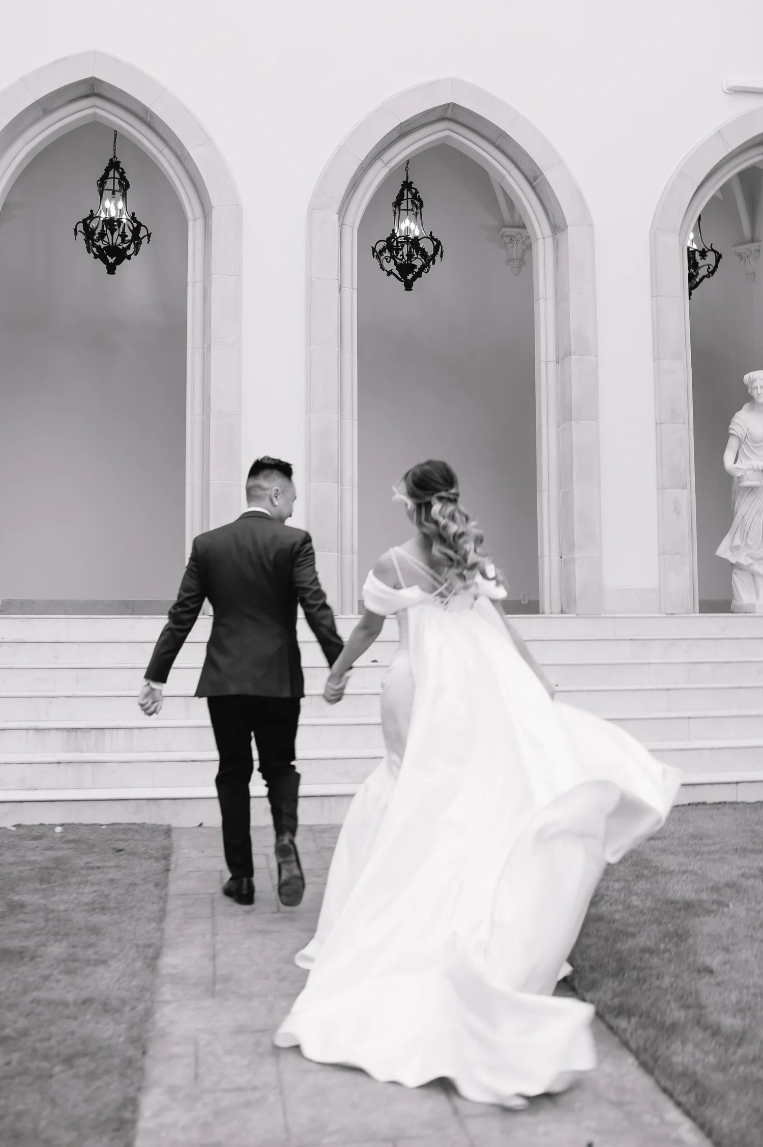 A romantic black and white photo of a couple running through an arched corridor at their wedding.