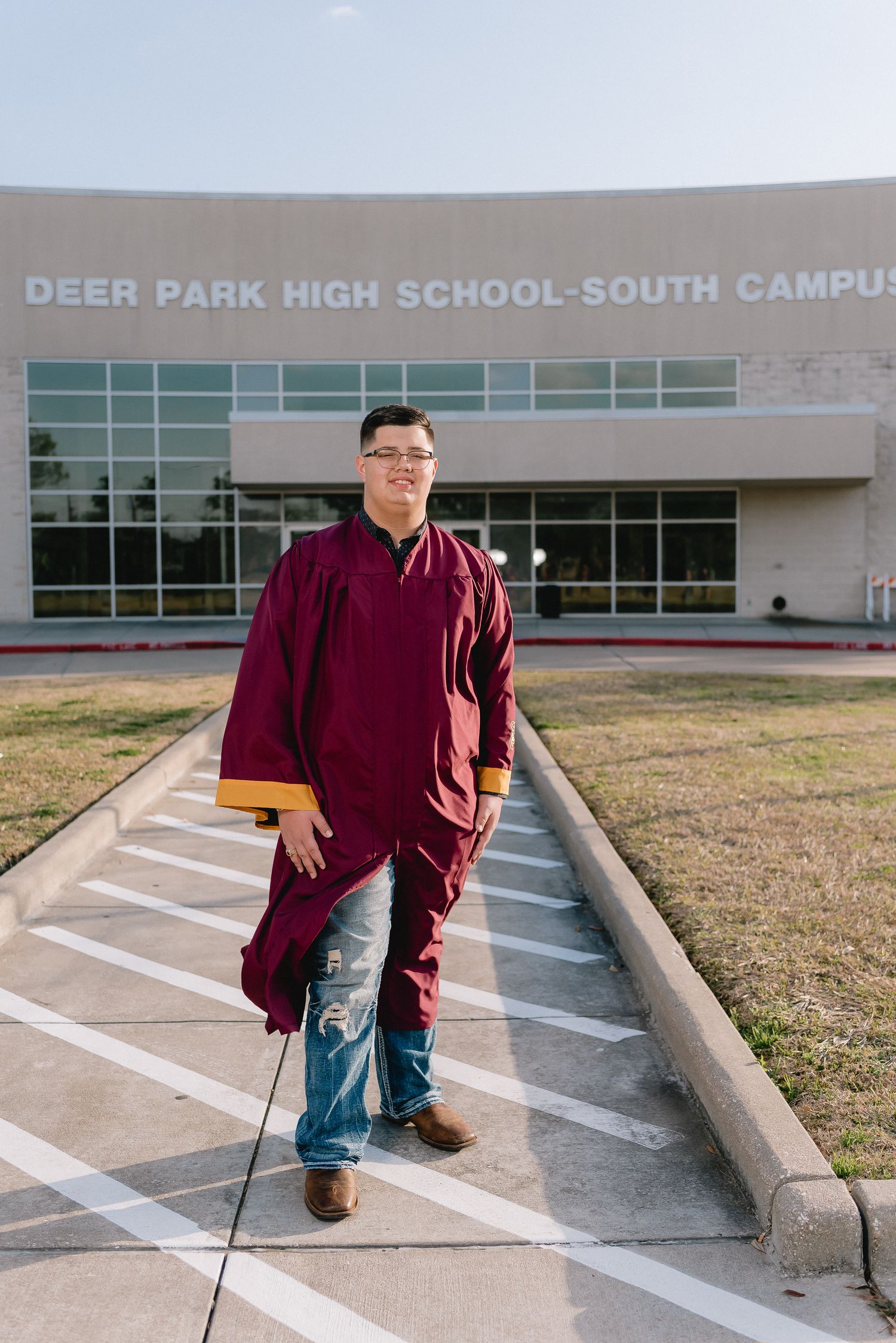 Graduate in maroon cap and gown standing in front of Deer Park High School-South Campus.