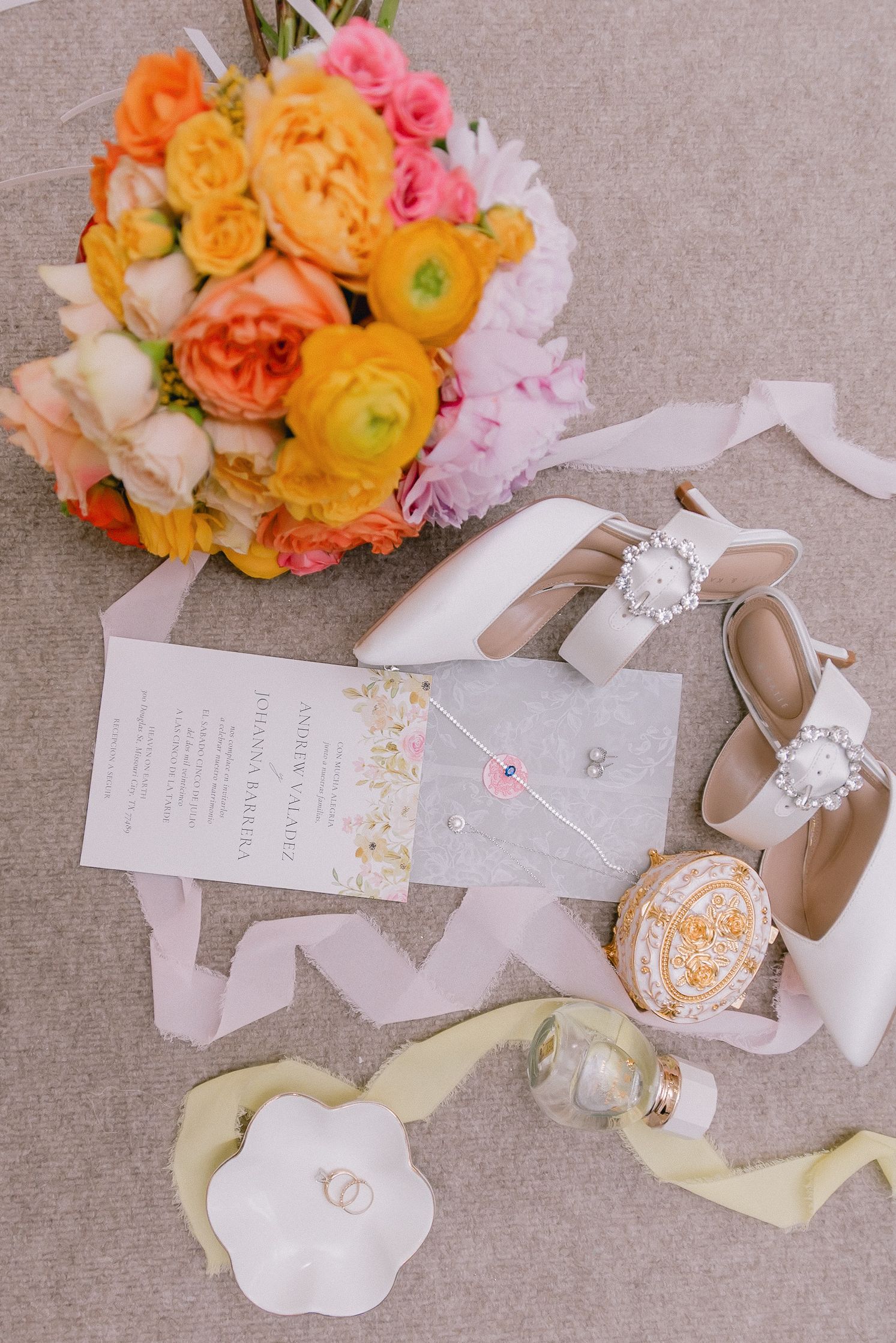 Wedding details flatlay with orange and pink flowers, invitations, and bridal shoes on grey background.