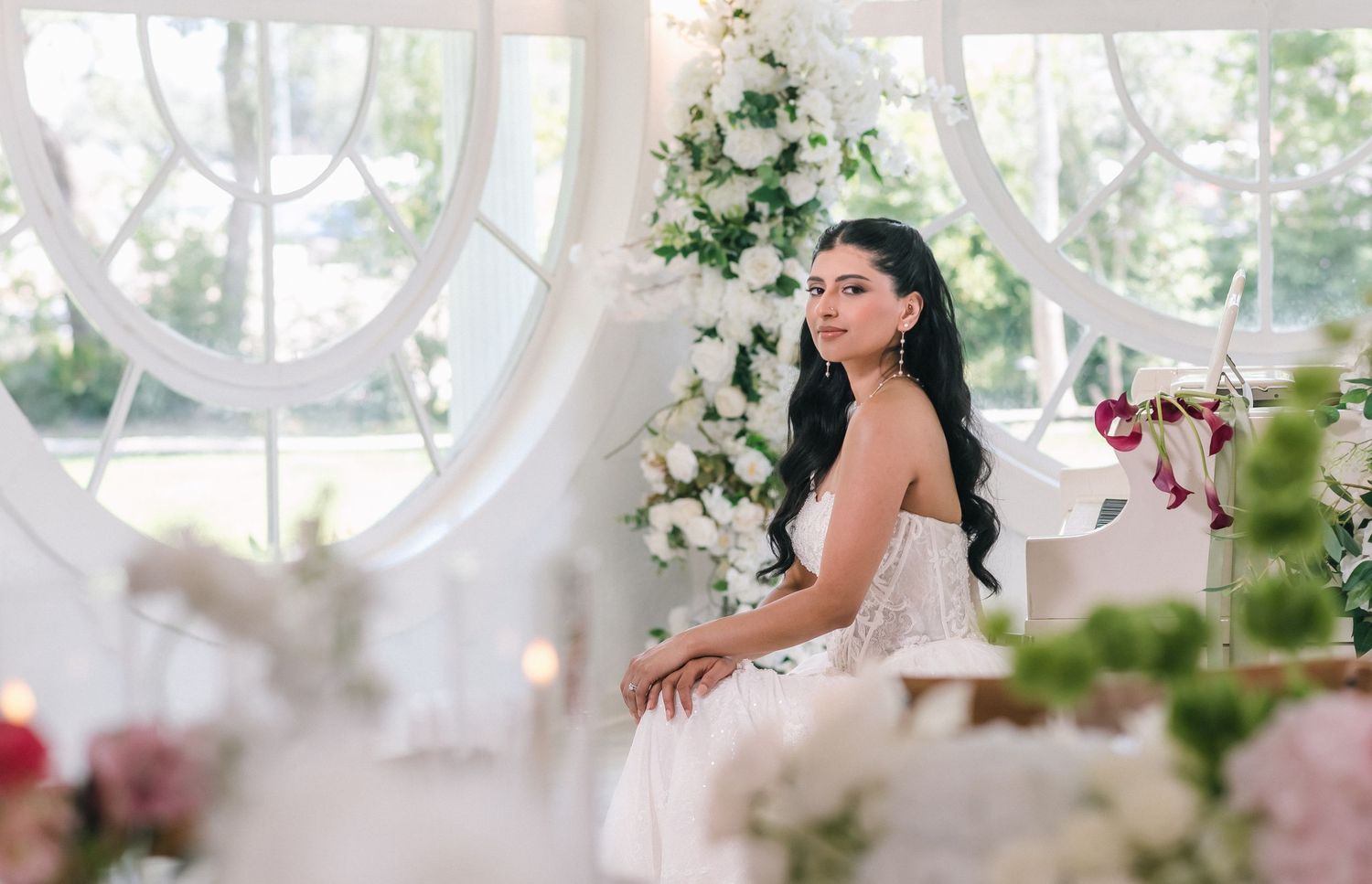 Elegant bride in white gown seated among floral arrangements in a bright, romantic wedding venue.