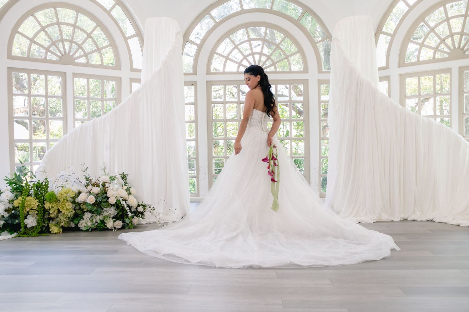Bride in elegant white ball gown with long train stands before arched windows with floral arrangements.