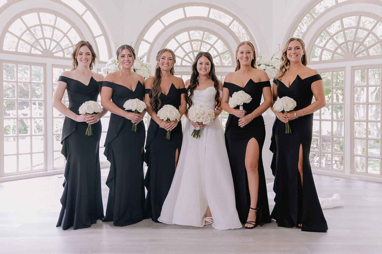 Wedding party in black and white attire poses elegantly in a bright venue with arched windows.