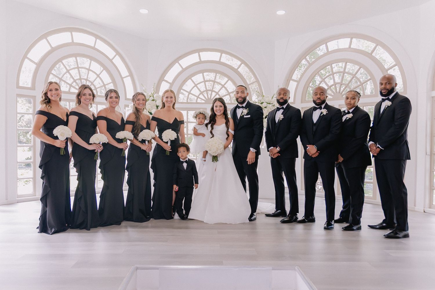 Wedding party lined up in formal black attire against white walls and arched windows.