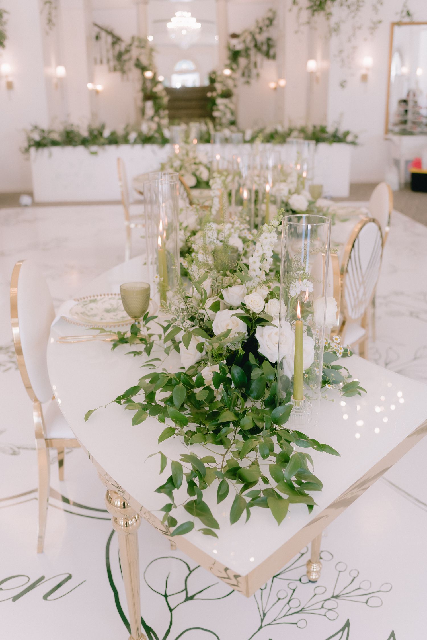 Elegant white wedding reception tables with green foliage centerpieces and gold accents in a bright venue.