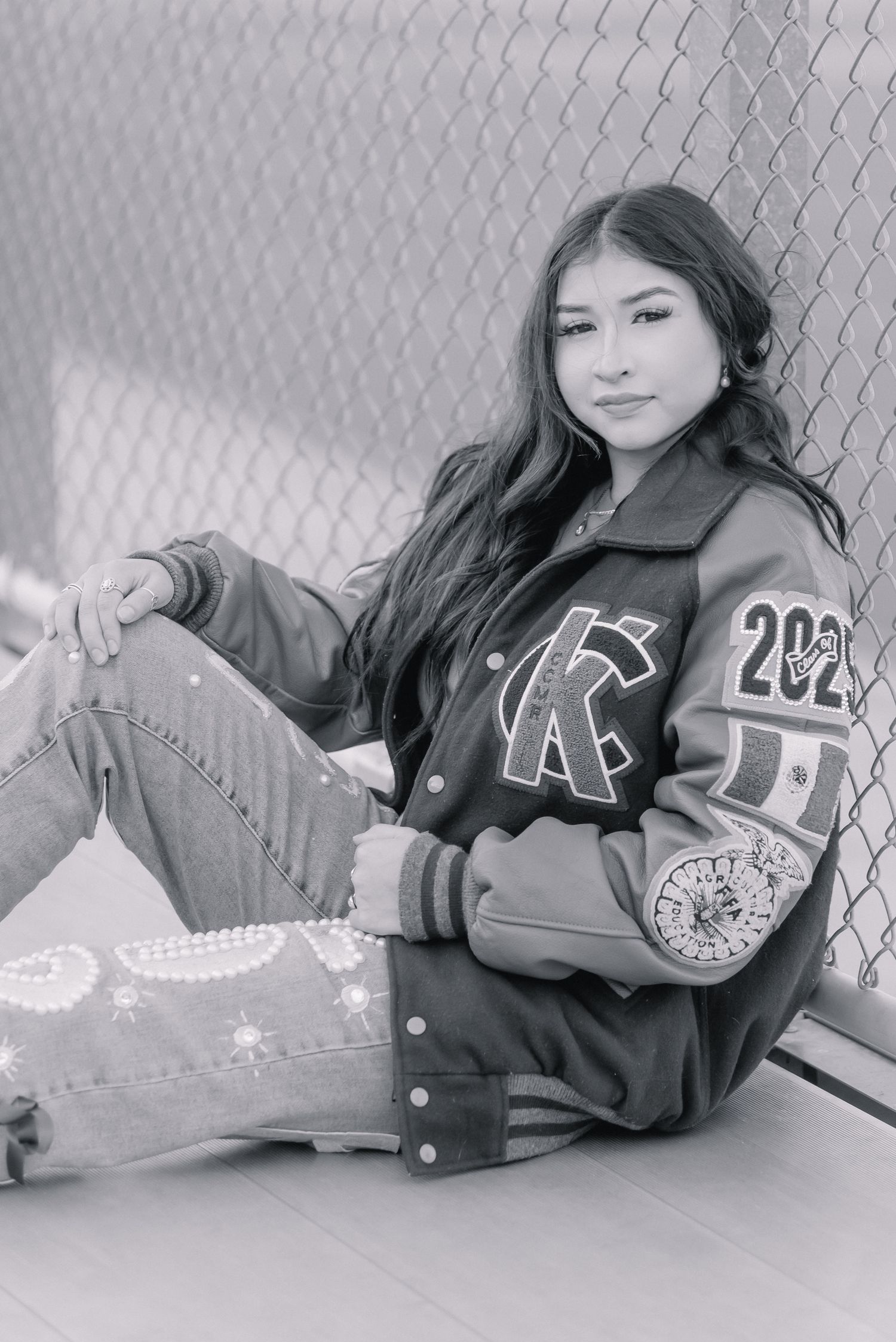 Black and white senior portrait in varsity jacket against chain link fence.