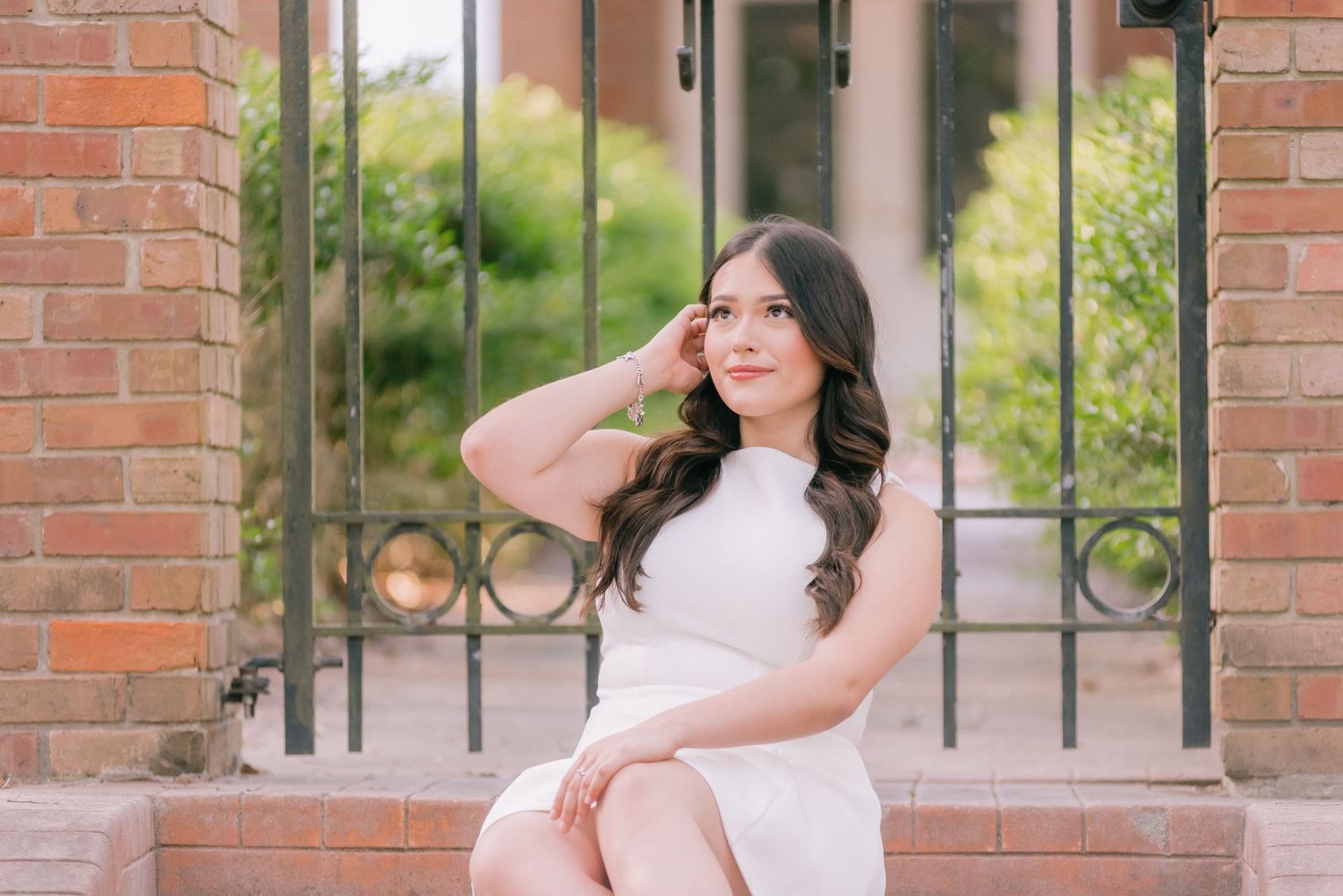 Elegant portrait in white dress seated by brick wall and iron gate.