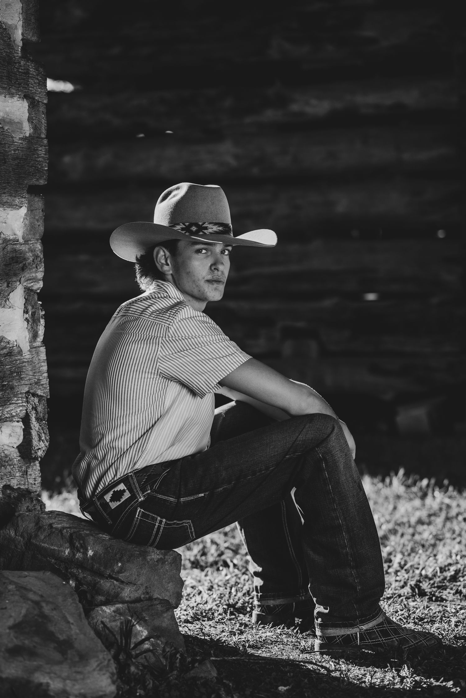 Black and white portrait of person in cowboy hat sitting against stone wall.