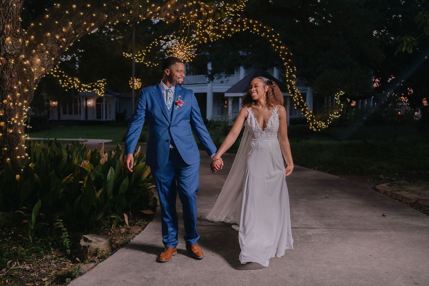 A couple walks hand in hand along a path illuminated by romantic string lights at dusk.