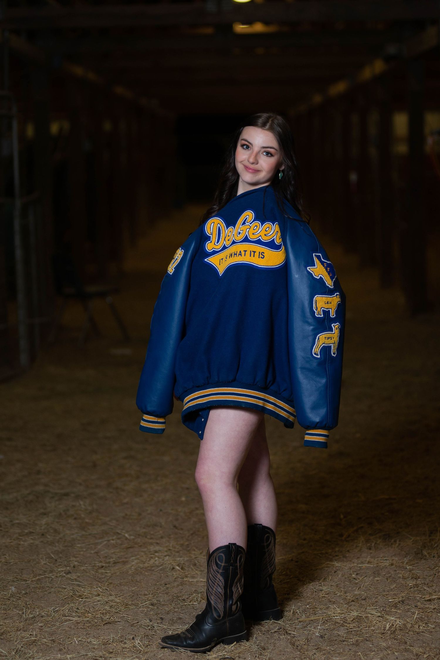 A blue and gold varsity jacket styled with cowboy boots in a rustic barn setting.