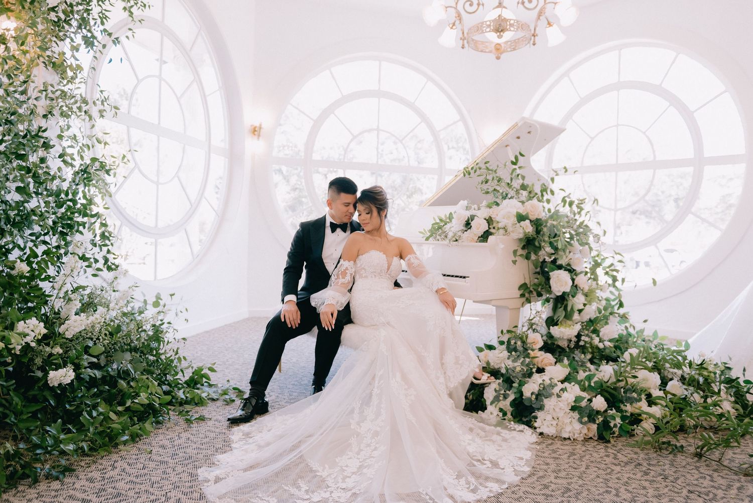 Bride and groom pose elegantly by a white grand piano adorned with white florals in a bright, romantic wedding venue.