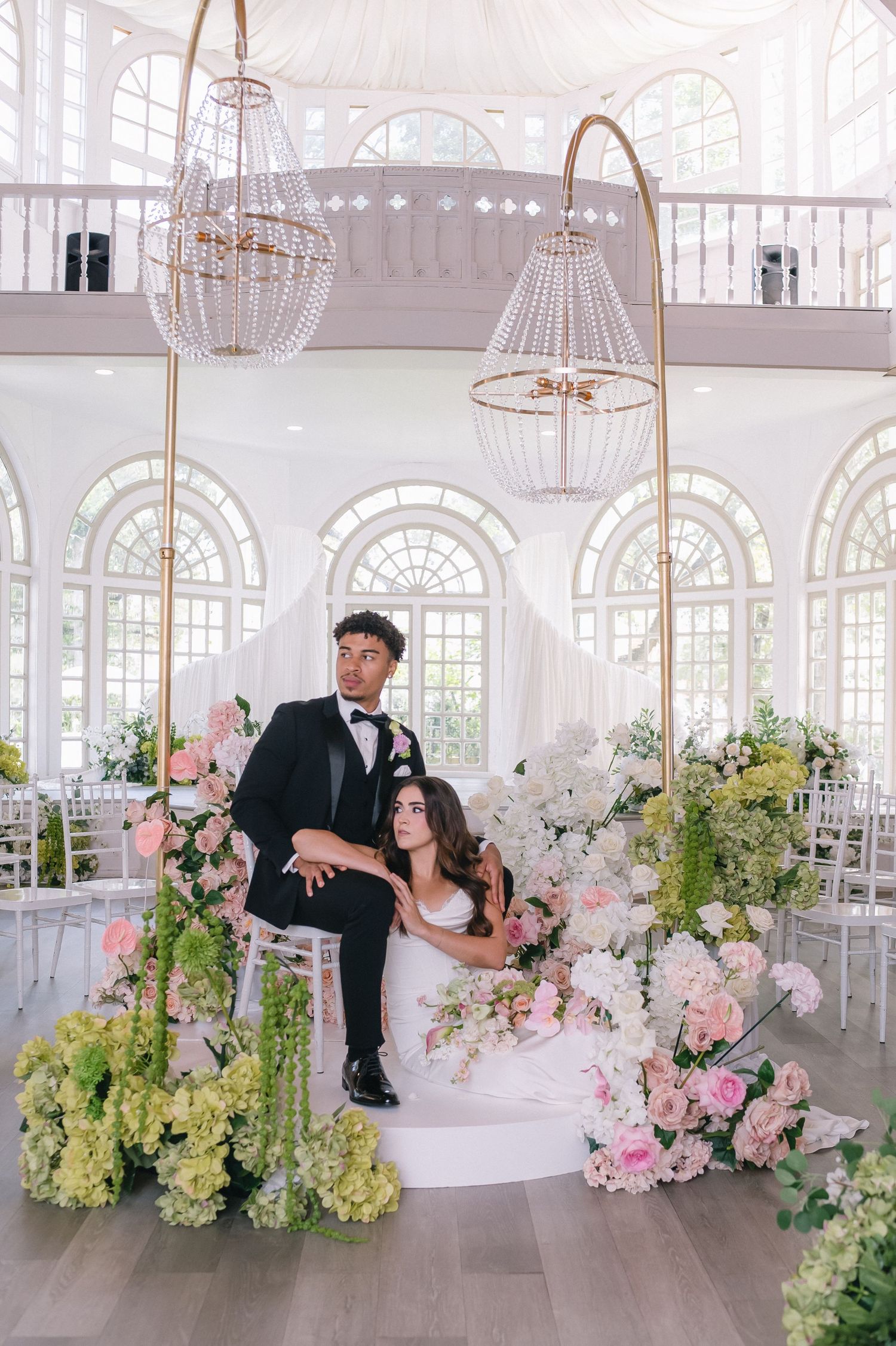 Bride and groom posing under gold arch with pink floral arrangements in elegant white wedding venue.