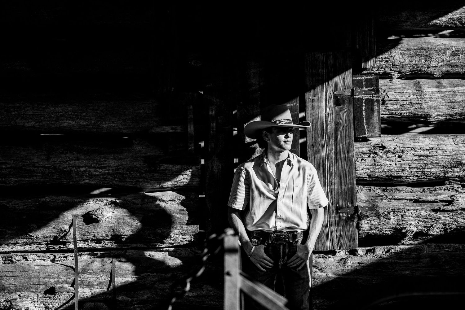 Black and white portrait of person in western wear against wooden cabin backdrop.