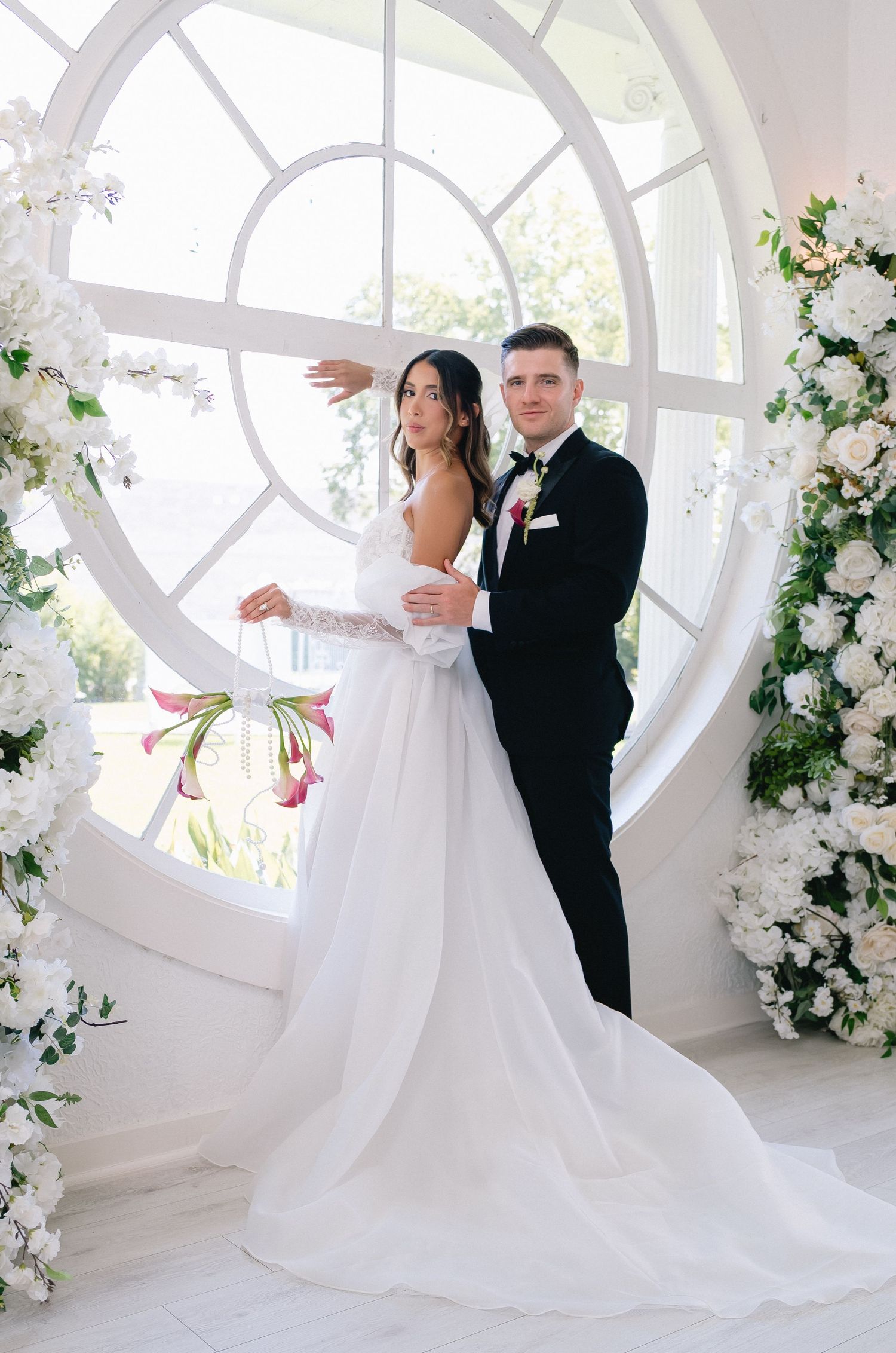 Bride in white gown and groom in black tuxedo pose before a circular window adorned with white floral arrangements.