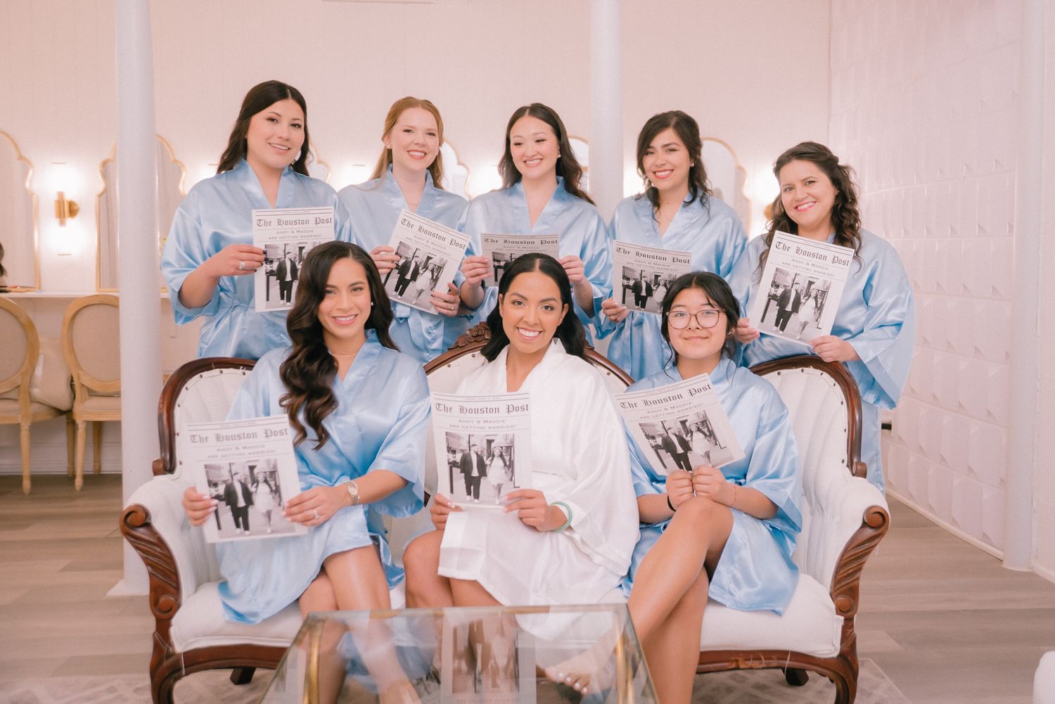 Group photo of bridesmaids wearing matching light blue robes in a bright indoor setting.