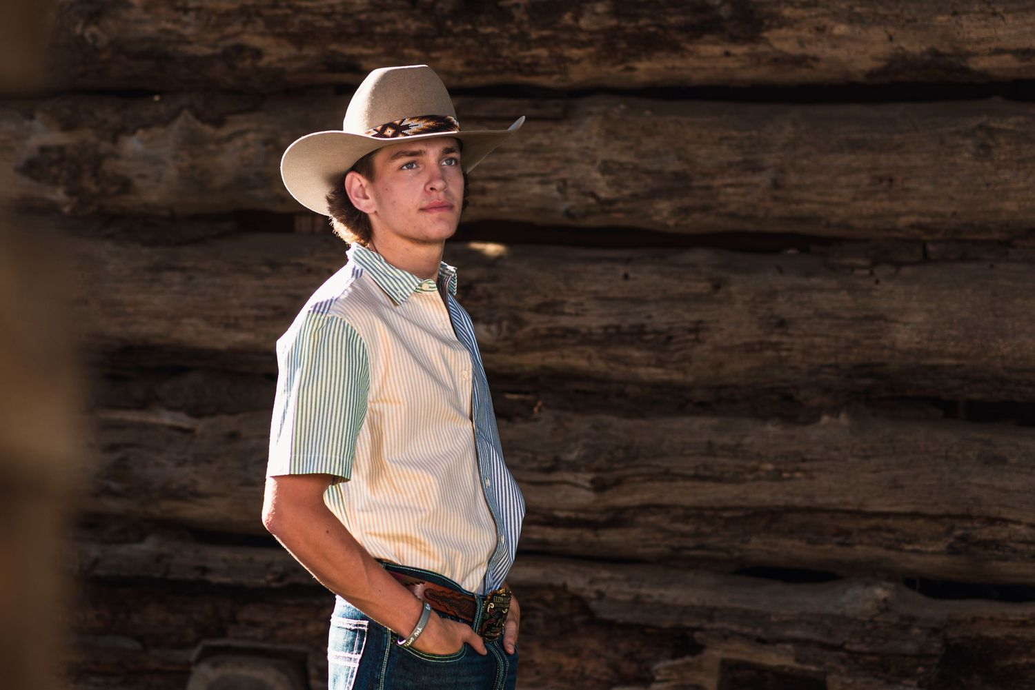 Western-style portrait against rustic log cabin wall featuring cowboy hat and casual attire.