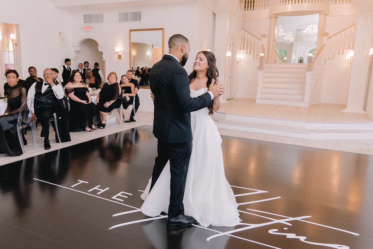 Couple shares first dance on patterned dance floor in elegant white ballroom.