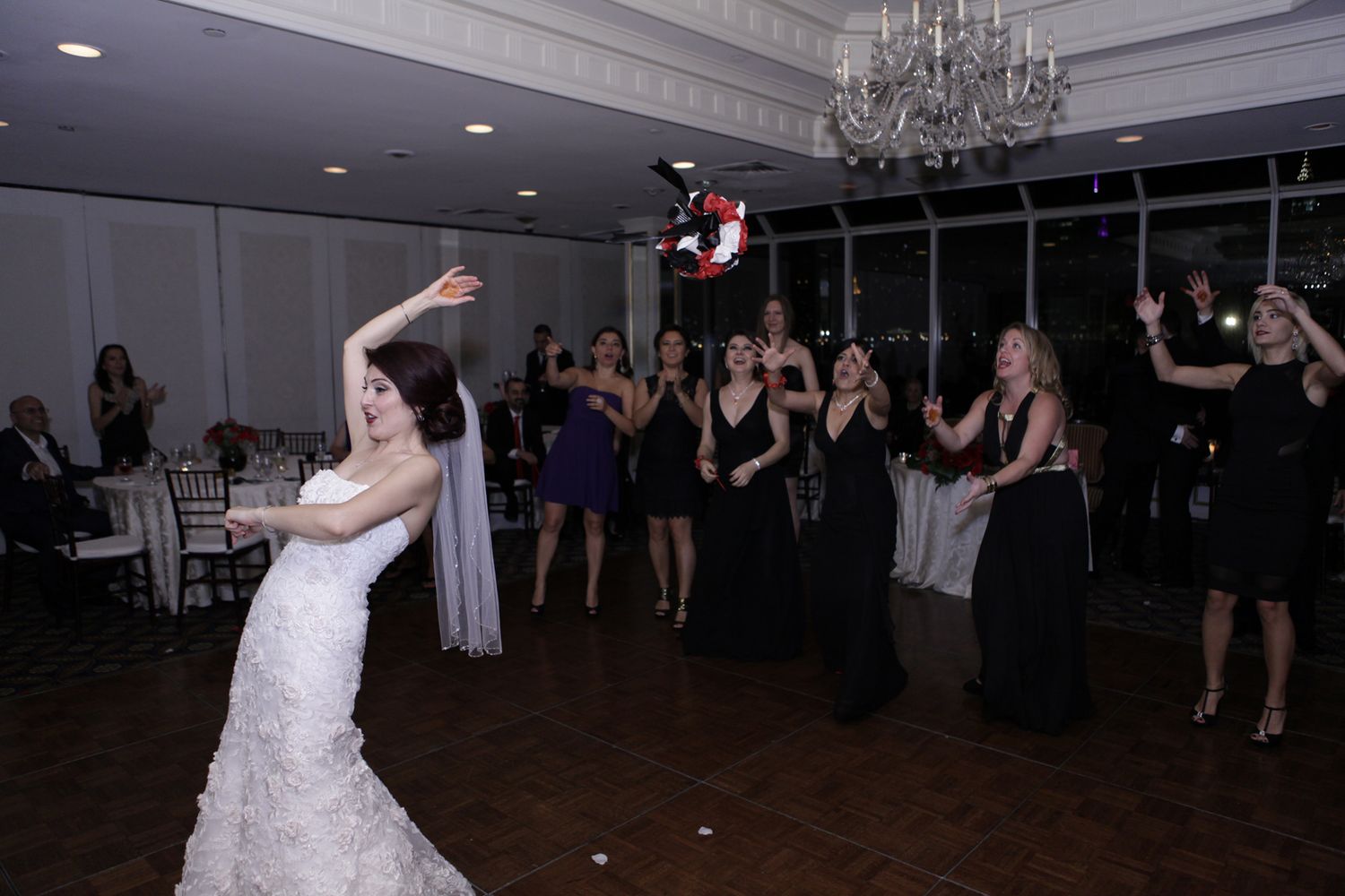 Wedding guests in black dresses watch as bride throws bouquet during reception.