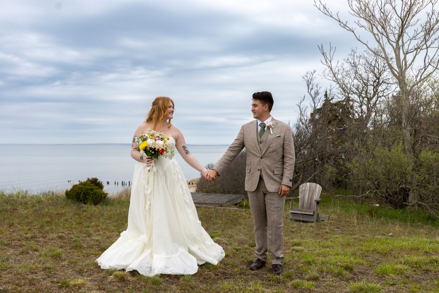 Bride in white gown holding colorful bouquet and groom in tan suit hold hands outdoors near seaside on cloudy wedding day.