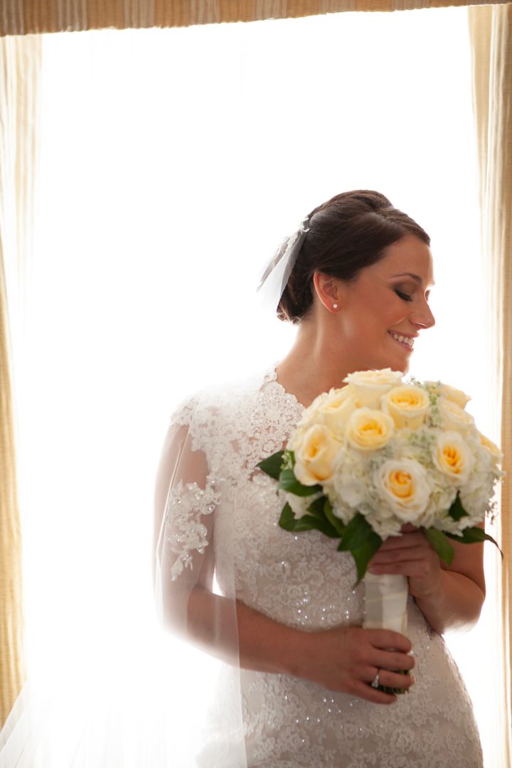 Smiling bride in delicate lace gown holds yellow rose bouquet.