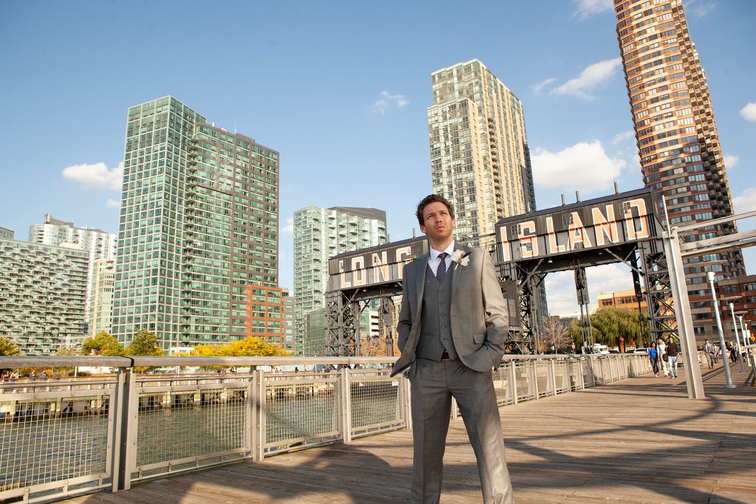 Man in suit poses on waterfront with Long Island City skyline and Pepsi Cola sign in background.