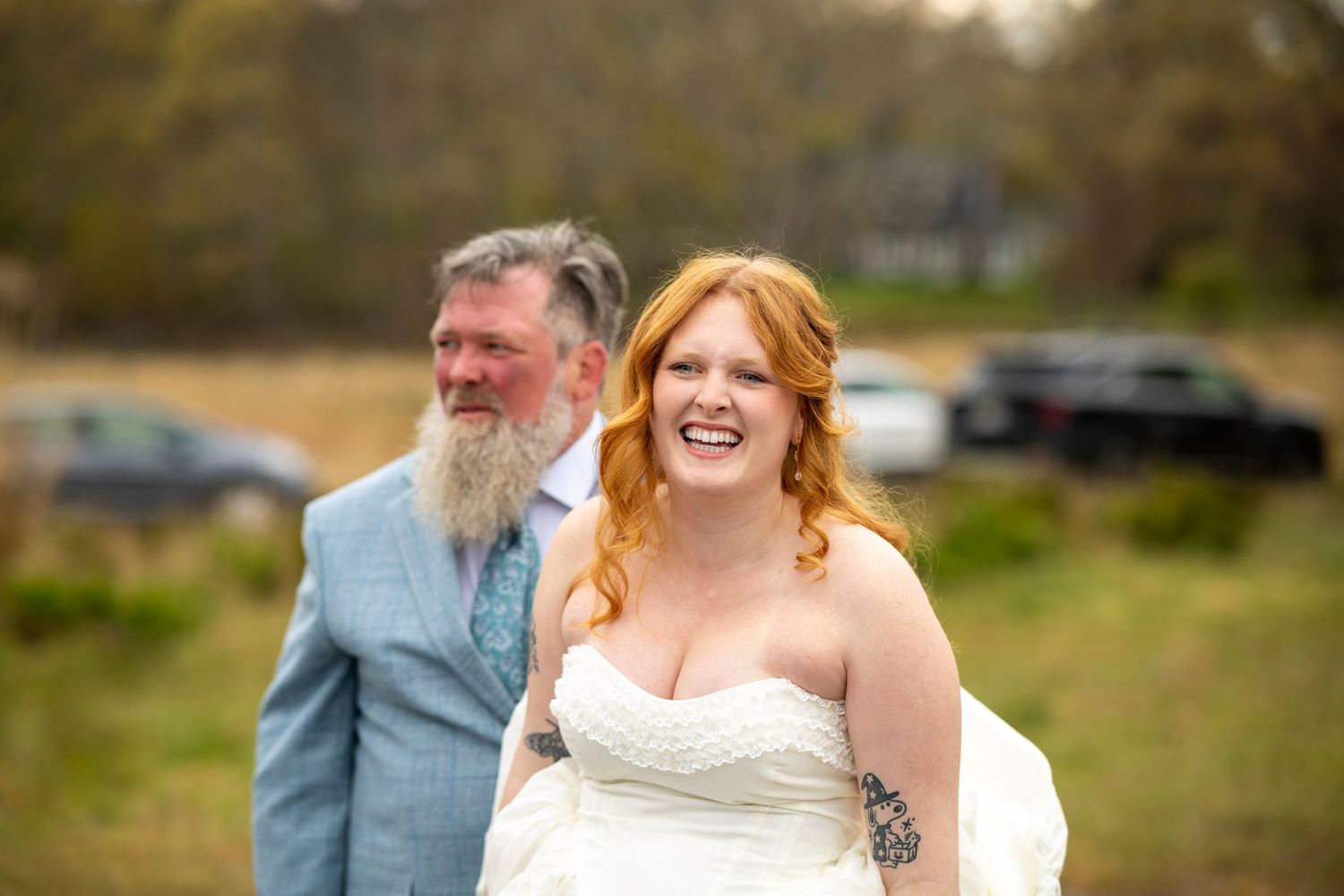 Bride in white strapless gown laughing joyfully outdoors, bearded groom in light blue suit standing behind her.