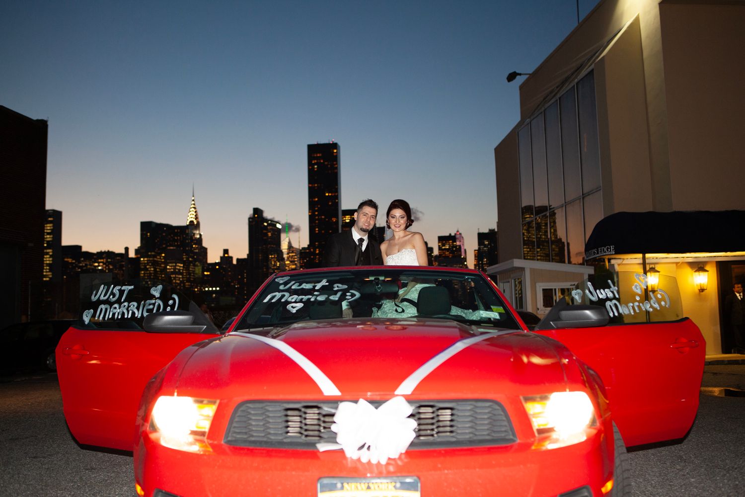 Wedding couple standing through sunroof of red decorated Mustang convertible at night.