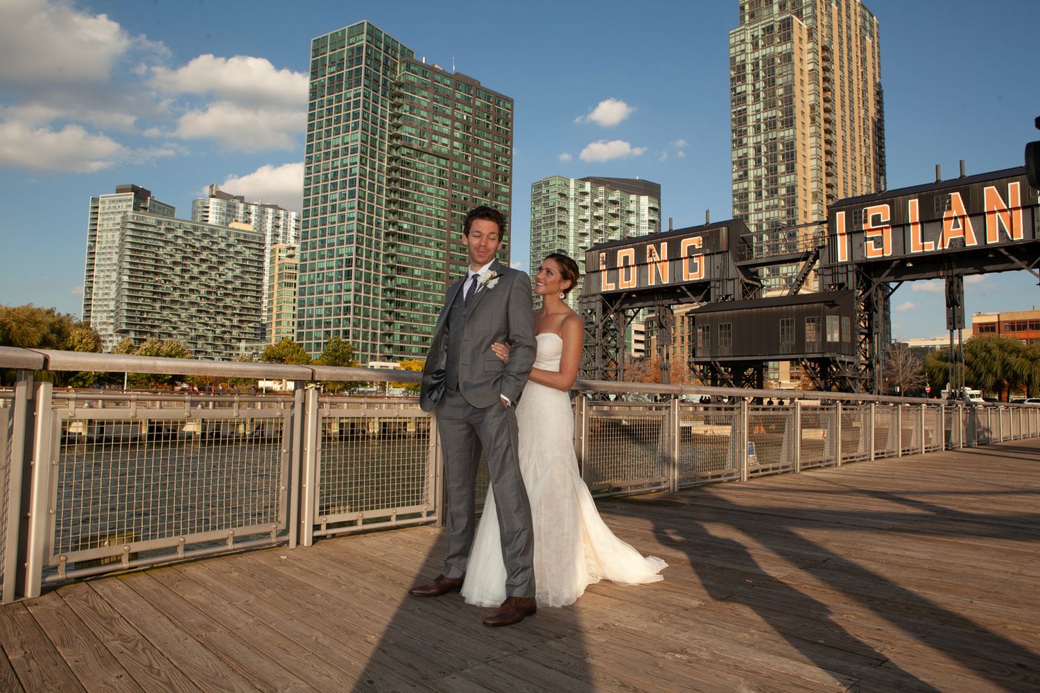 A wedding couple poses on a pier with modern city buildings in the background.