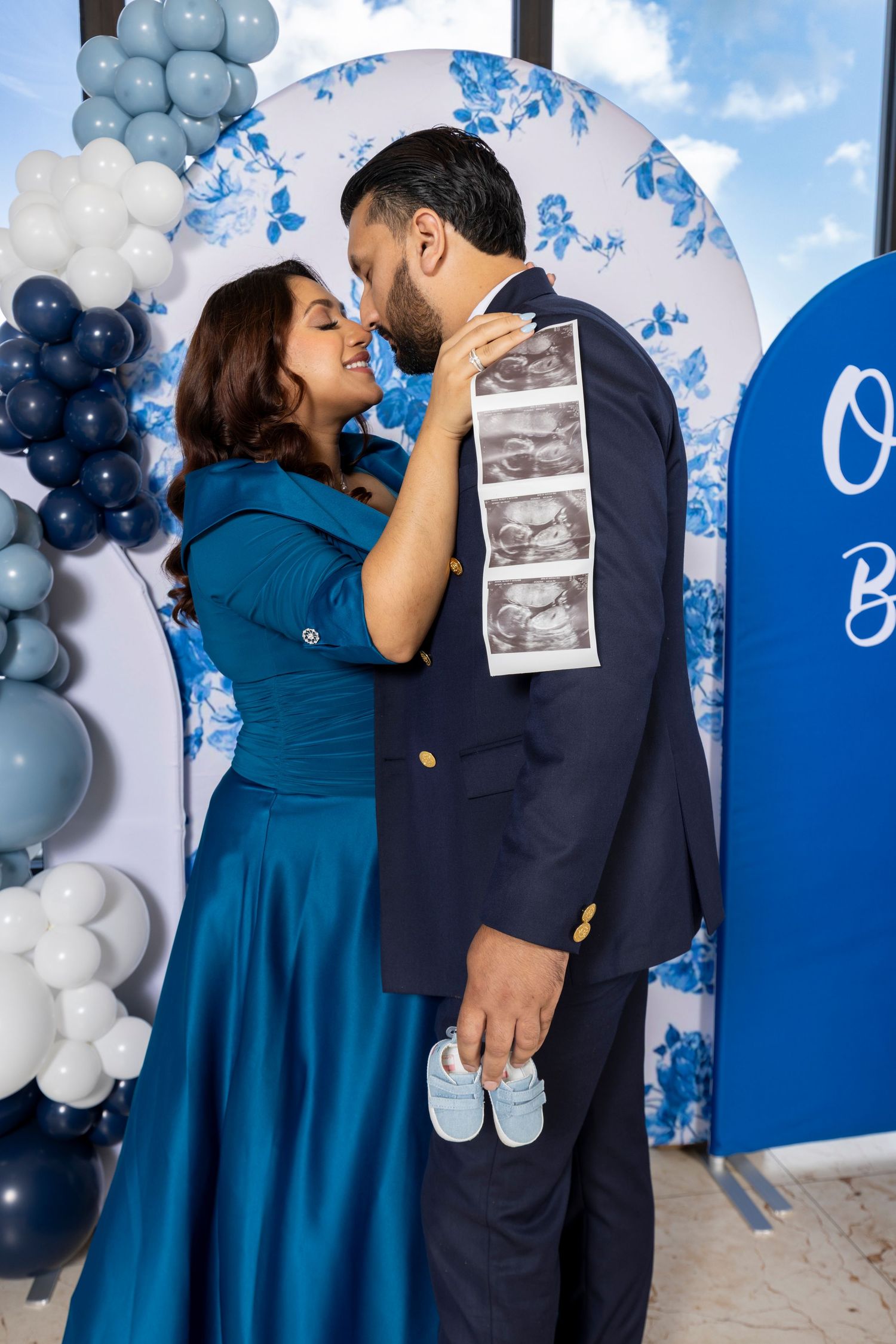 A couple embraces in front of a blue and white balloon arch with ultrasound photos displayed during a gender reveal party.