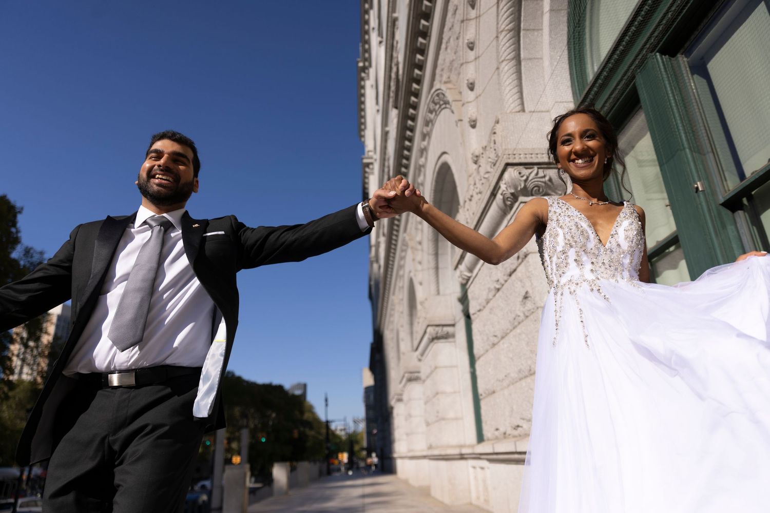 A bride in a white lace gown and groom in a black suit hold hands joyfully outside an elegant stone building.