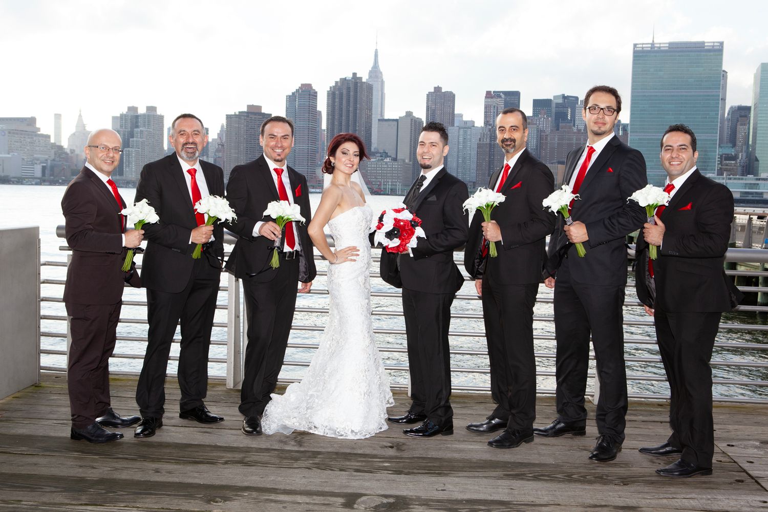 Wedding party in formal attire poses with bouquets against NYC waterfront.