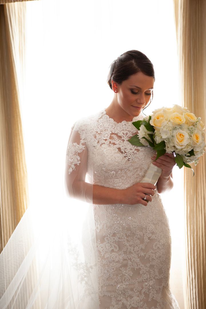 Elegant bride in lace dress holds yellow rose bouquet near window.