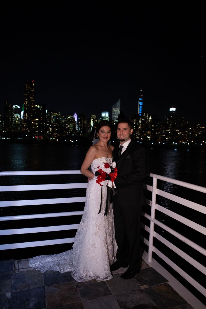 Formal couple poses on waterfront deck with illuminated city skyline in background.