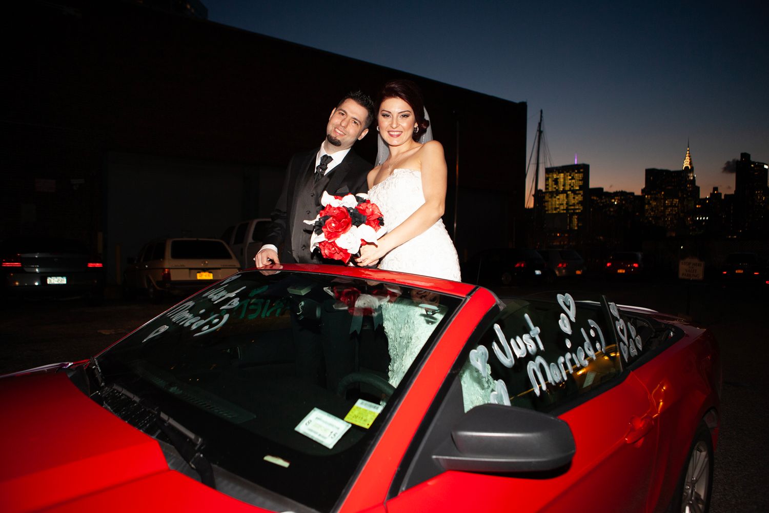 Wedding couple standing through sunroof of red convertible with city lights in background.