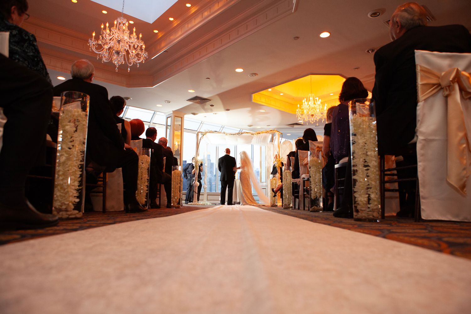 Elegant wedding ceremony venue with crystal chandelier and guests lining the aisle in formal attire.