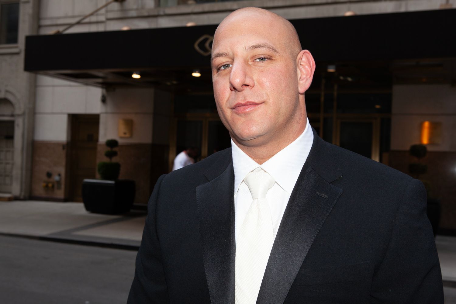 Man in formal black tuxedo and white shirt poses outdoors near columns.