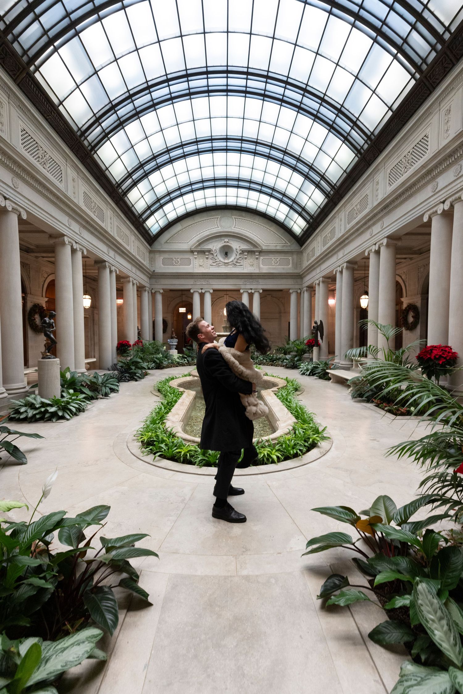 A person spins joyfully in a grand indoor courtyard with columns, skylights, and lush greenery at The Frick Collection.