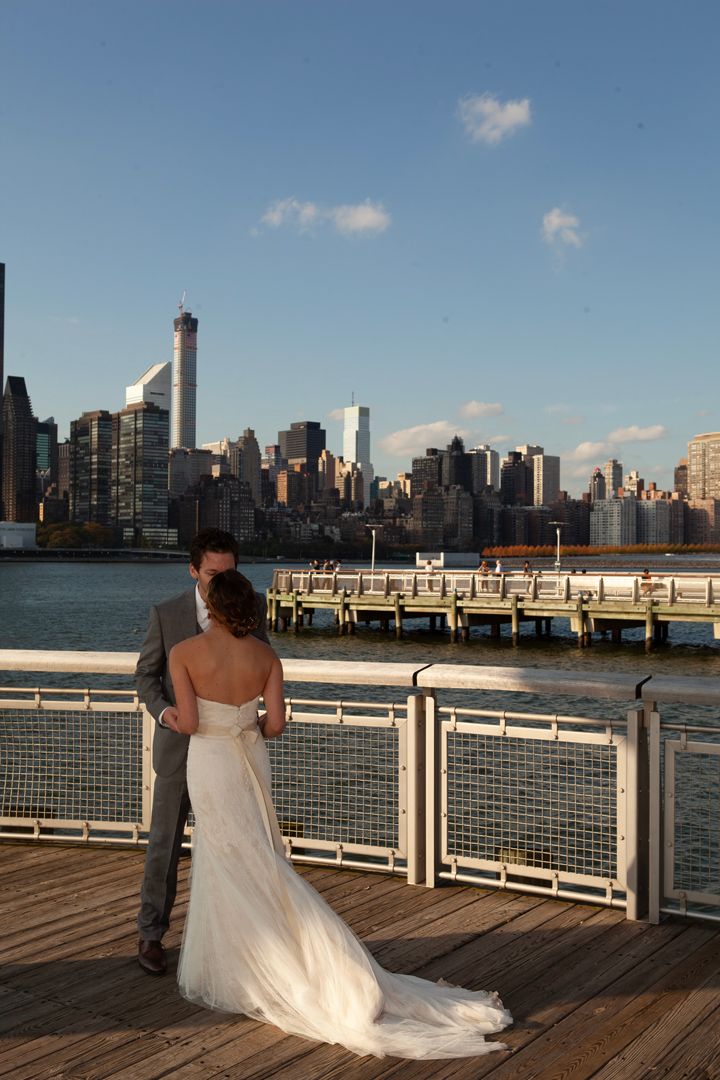 A wedding couple embraces on a waterfront pier with the NYC skyline behind them.