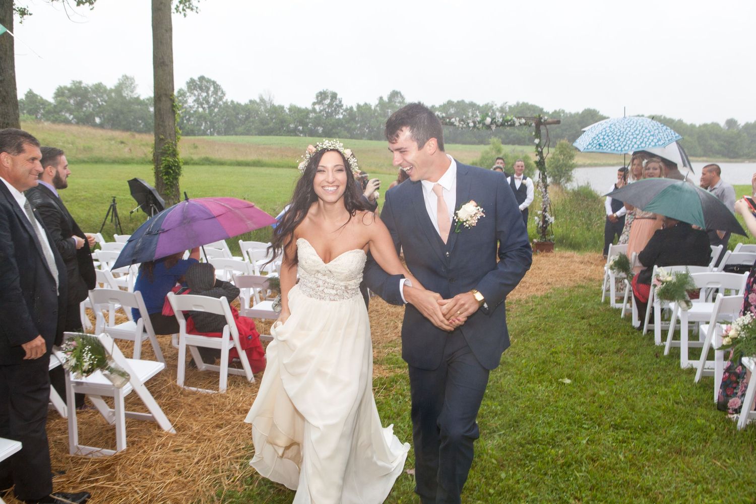A joyful couple runs through their wedding ceremony aisle while guests hold umbrellas in the rain.