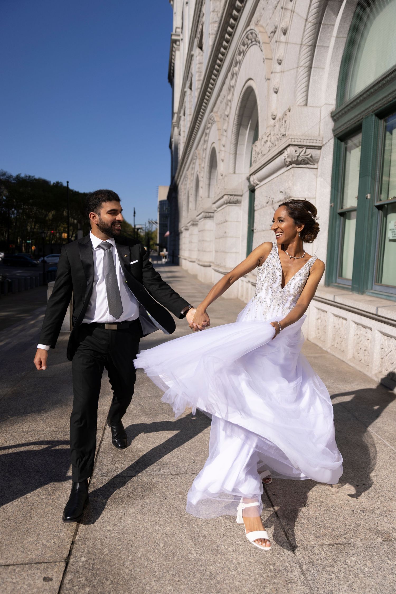 A couple in wedding attire dance joyfully outside a historic building with ornate architecture on a sunny day.